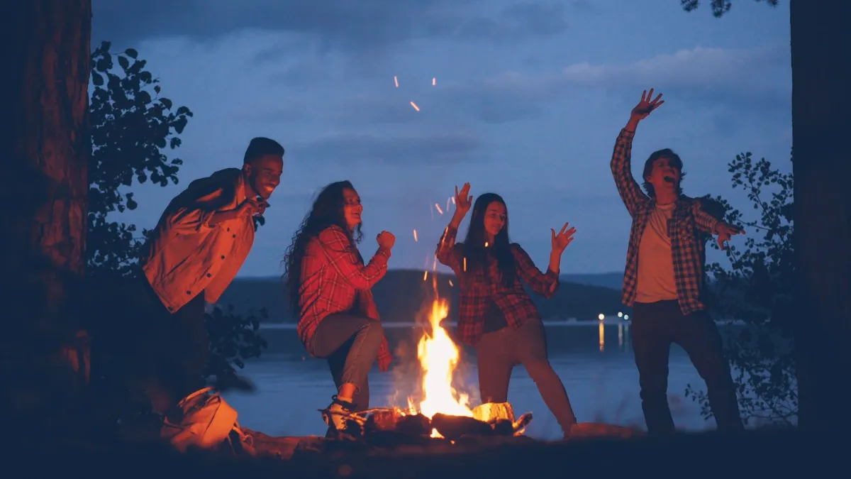 Amigos bailando alrededor de una hoguera de camping al anochecer